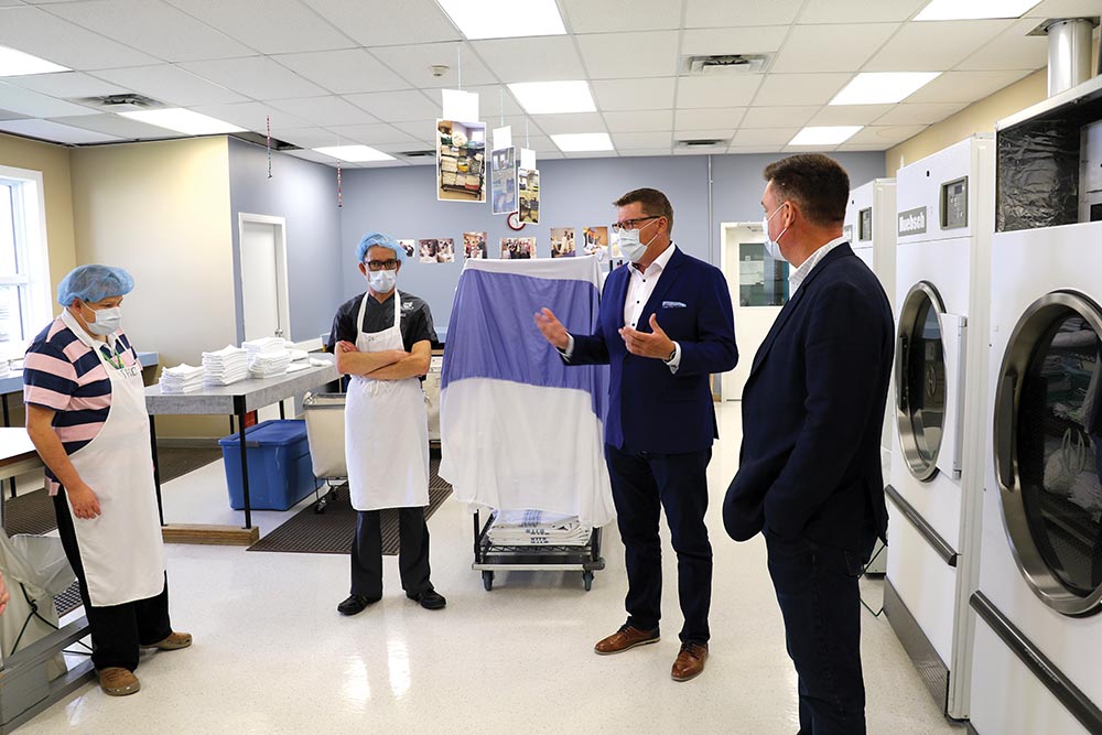 Saskatchewan Premier Scott Moe and Moosomin MLA Steven Bonk visit with Moosomin Kin-Ability Centre clients Ryan Bender and Barry Gessell while learning about the hospital laundry services offered at the Kin-Ability Centre.